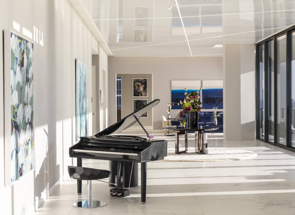 Sunlit hallway in a One Thousand Museum residence featuring a black grand piano, abstract artwork, and panoramic glass views.