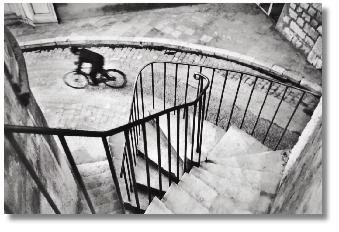 A cinematic black-and-white photo of a cyclist rounding a curve, capturing fleeting motion from above—a tribute to French humanist photography and the poetic realism of mid-century Paris.
