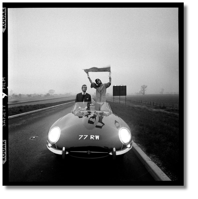 A stylish couple is captured on a foggy road with a Jaguar E-Type; the woman stands up in the car holding a map above her head.