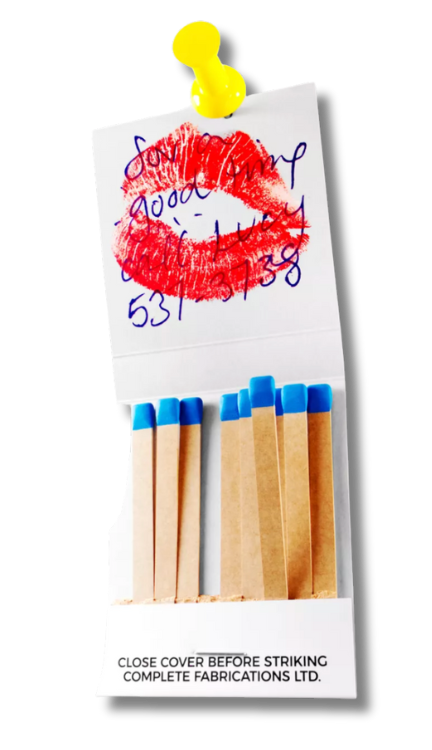 A matchbook with blue-tipped matches and a handwritten note above a lipstick kiss, pinned with a yellow thumbtack.