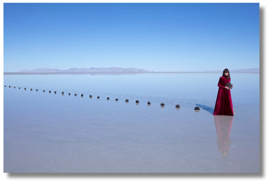 A woman in a rich red gown stands in a serene salt flat with distant mountains, holding a volcanic rock.