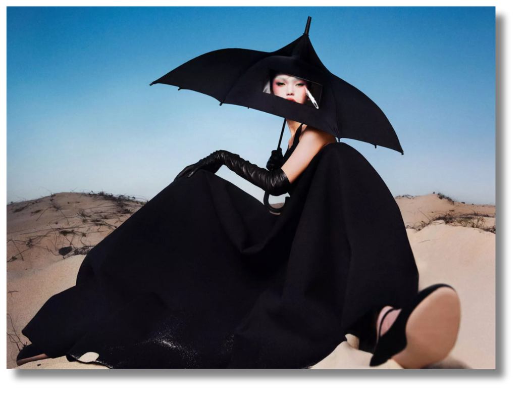 A high-fashion portrait in a desert setting, where a model in dramatic black attire holds an umbrella against the sky.