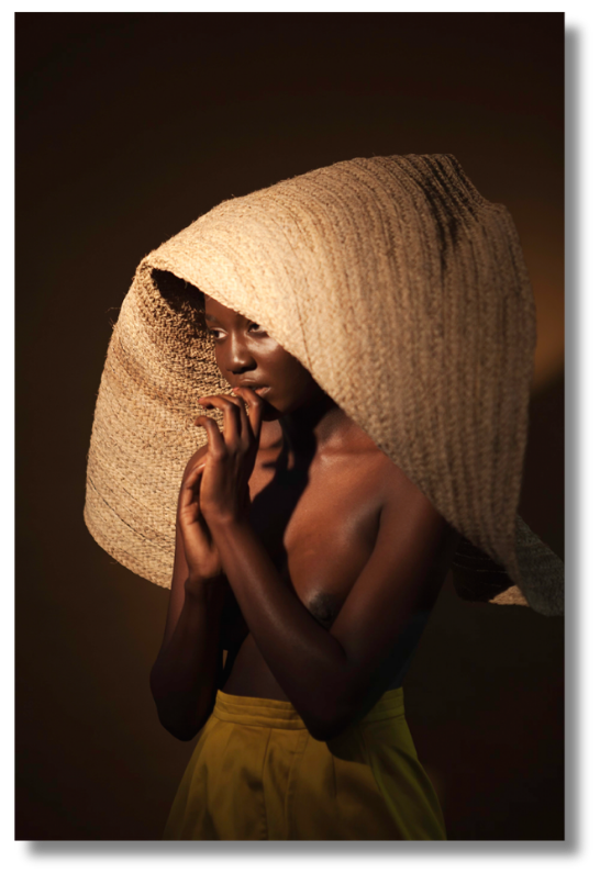 Figure wearing oversized woven natural fiber headpiece under warm directional lighting. Contemporary piece from the electric horizon art collection highlighting texture, shadow, and sculptural elegance.